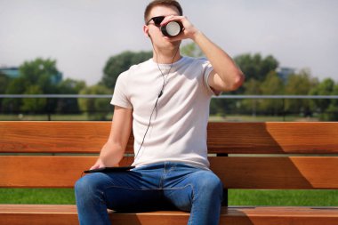 Gitmek için yakışıklı çocuk tablet ve kulaklık kullanarak, bir bankta oturan ve kahve içme güneş gözlüğü takıyor. Yakışıklı öğrenci sosyal medyada dalmış. Arka plan manzara Köprüsü, nehir ve yeşil çimen.