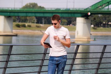 Tablet ve kulaklık kullanarak çit yaslanmış adam onun çevresi nehir ve eski çelik, yeşil renkli Köprüsü tarafından zevk. Şehir, şehir çevre ve sahne. Açık Şehir içinde vaha.