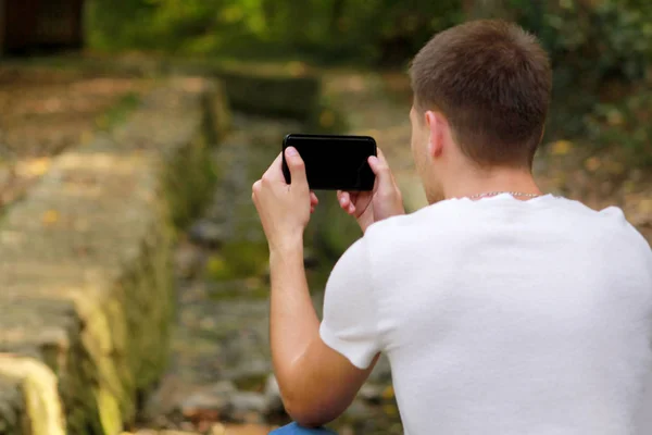 Young man taking pictures using camera his smart phone in nature, park and forest, a small river, a stream. Hand male holding smart phone taking picture of beautiful landscape scenery background.