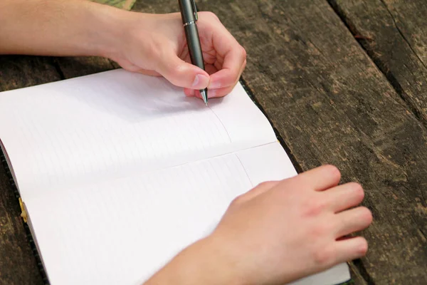 Young handsome guy sitting at wooden table, writing a book, doing homework, taking notes, learning, contemplating and writing down his thoughts and lessons while enjoying the outdoors in forest.