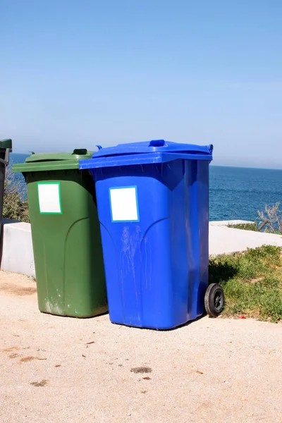 Trash can, garbage bin, recycling bin in tourist complex by sea, side of road waiting to be picked up by garbage truck. Blue and green containers for waste sorting, sort garbage for paper and glass.