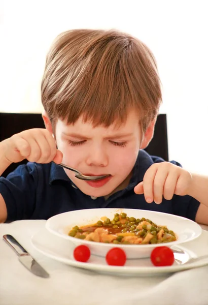 Criança com fome sentada na cadeira à mesa na cozinha e comendo com colher de ervilhas cozidas com tomate. Crianças refeição e dieta saudável conceito de comida. Menino feliz e bonito desfrutando em bom almoço em casa . — Fotografia de Stock