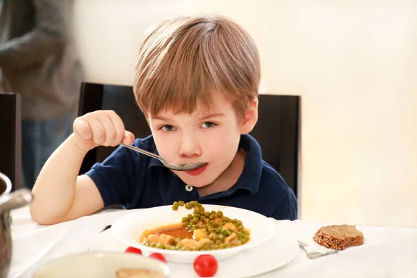 Hungry child sitting in chair at table in kitchen and eating with spoon of cooked peas with tomato. Kids meal and healthy diet food concept. Happy and cute little boy enjoying in good lunch at home.