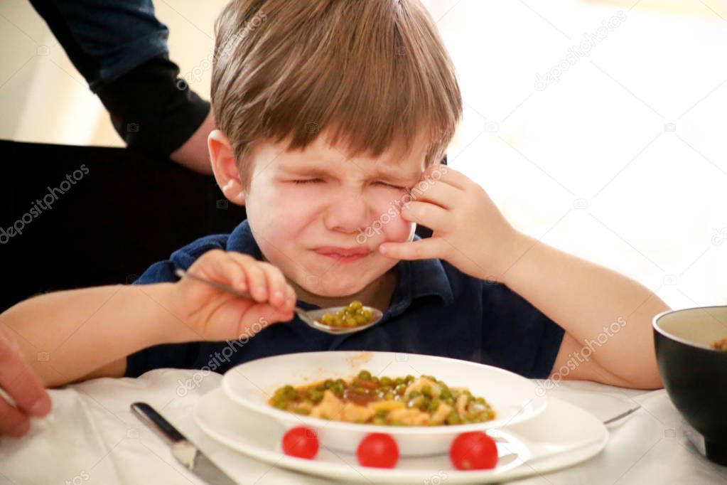 Hungry child sitting in chair at table in kitchen and despises, disapproves of hot meal, because he does not like lunch. Kid is having of cooked peas with tomato for lunch. Healthy diet food concept.