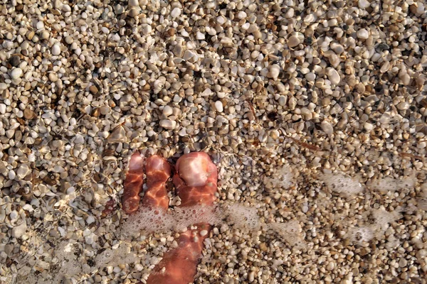 Top view of man standing bare feet on beach. Texture of bottom, leg and foot of man drowning with sweeping colorful stone below on rocky stones beach in sea with waves and foam, water ripples of sea. — Stock Photo, Image