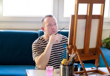 Erkek sanatçı ressam sanat boya atölyesinde ahşap çizim tahtası şüskül üzerinde tuval ile atölyede çalışıyor. Boya fırçası ile sanatçının portresi yağlı boyalar ile tuval üzerine boyama olduğunu. Yaratıcılık kavramı.