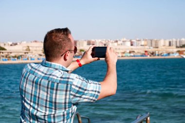 Sahil iskelesindeki turist ve gezgin, deniz kıyısı, çevre ve doğa manzarasına ait akıllı telefonunu kullanarak fotoğraf çekiyor. Plaj platformunda otel tatilde güneş gözlüğü ile Adam.