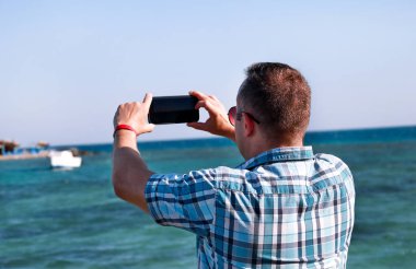 Sahil iskelesindeki turist ve gezgin, deniz kıyısı, çevre ve doğa manzarasına ait akıllı telefonunu kullanarak fotoğraf çekiyor. Plaj platformunda otel tatilde güneş gözlüğü ile Adam.