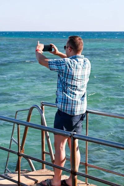 Turista y viajero en el muelle de la playa está tomando fotos utilizando la cámara de su teléfono inteligente de la costa del mar, el medio ambiente y el paisaje natural. Hombre con gafas de sol de vacaciones en el complejo hotelero en la plataforma de playa . —  Fotos de Stock