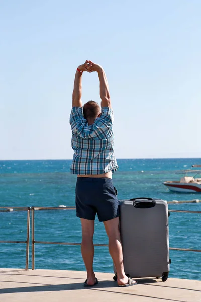 Turista y viajero está parado en la playa del complejo hotelero con maleta viajada, disfruta de vista al mar, paisaje natural, panorama, horizonte y descanso bajo el sol. Hombre guapo de vacaciones en verano . —  Fotos de Stock