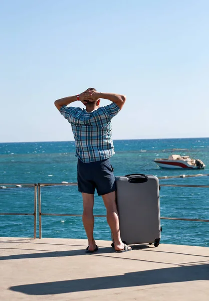 Turist ve gezgin seyahat bavul ile otel tesisi plajda durmak, manzara deniz ortamı, doğa manzara, panorama, ufuk ve güneşte dinlenme sahiptir. Yakışıklı adam yaz tatilde. — Stok fotoğraf