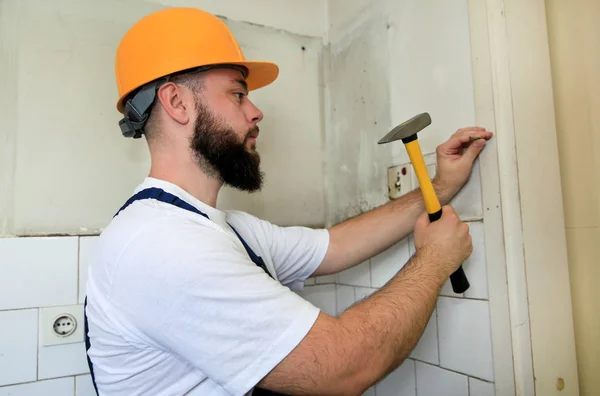 Trabajador de la construcción y manitas que trabajan en trabajos de renovación. Constructor con martillo amarillo golpea y clava un clavo en la pared de madera de la puerta de la cocina en el sitio de construcción. Concepto de renovación de apartamentos . — Foto de Stock