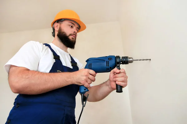 Construction worker and handyman works on renovation of apartment. Builder with blue electric drill drills a nail hole into wall of construction site. House renovation concept. Construction tools.