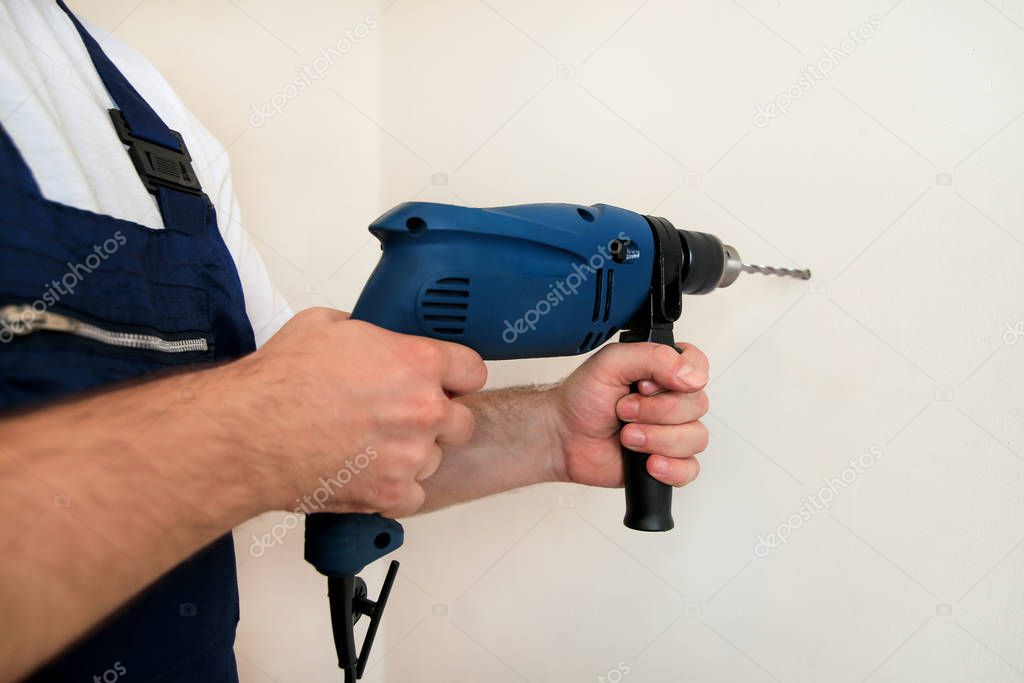 Construction worker and handyman works on renovation of apartment. Builder with blue electric drill drills a nail hole into wall of construction site. House renovation concept. Construction tools.
