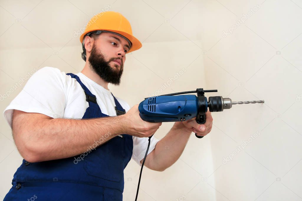 Construction worker and handyman works on renovation of apartment. Builder with blue electric drill drills a nail hole into wall of construction site. House renovation concept. Construction tools.