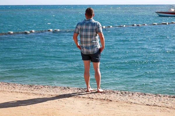 Retrato de turista y viajero está posando y observando en la playa de arena del complejo hotelero, disfruta de vistas al mar, paisaje natural, panorama, horizonte y descanso bajo el sol. Hombre de vacaciones en verano . —  Fotos de Stock