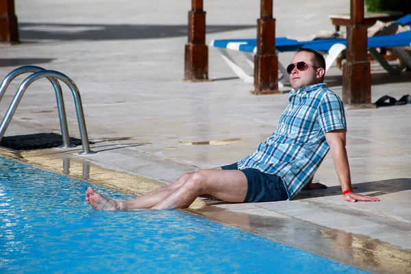 Turista y viajero está sentado, posando y acostado junto a la piscina del complejo hotelero, disfruta de vistas al medio ambiente y descansando en un día soleado. Un hombre guapo de vacaciones en verano. Concepto de vacaciones . —  Fotos de Stock