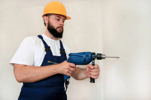 Construction worker and handyman works on renovation of apartment. Builder with blue electric drill drills a nail hole into wall of construction site. House renovation concept. Construction tools.