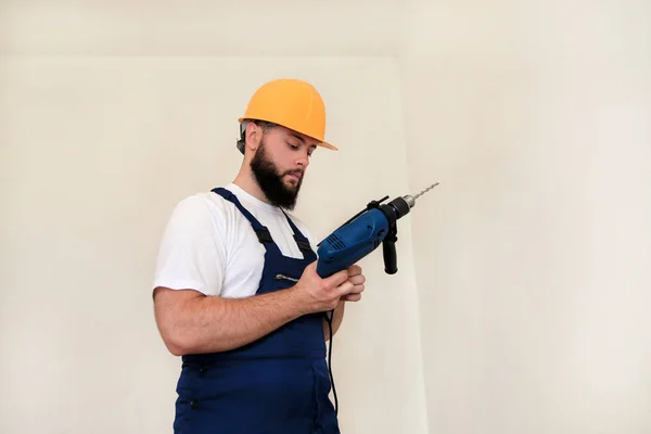 Construction worker and handyman works on renovation of apartment. Builder with blue electric drill drills a nail hole into wall of construction site. House renovation concept. Construction tools.