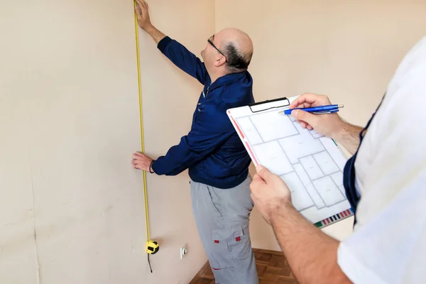 Ingenieur, aannemer en Project Manager met zijn collega, bouwvakker, klusjesman en bouwer is het meten van de muur van ruimte voor renovatie met behulp van meetlint en controle in appartement blauwdruk. — Stockfoto