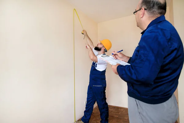 Ingeniero, contratista y director de proyecto con su colega, trabajador de la construcción, manitas y constructor está midiendo la pared de espacio para la renovación utilizando cinta métrica y comprobando en el plano del apartamento . — Foto de Stock