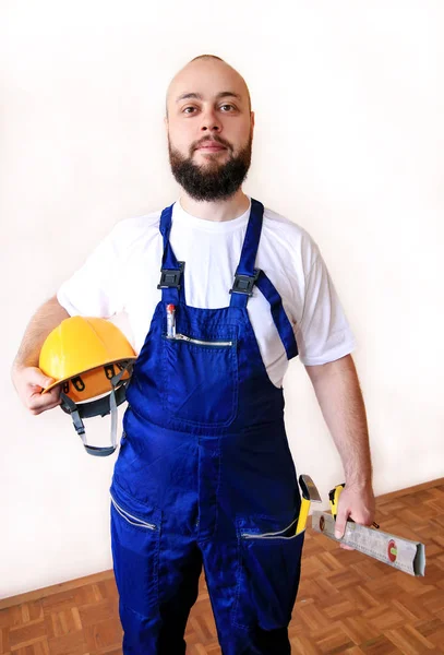 Construction worker at construction site with protective uniform, construction service tools and equipment, coverall and holds yellow helmet, hammer tool, measure tape, spirit level tool in hands.