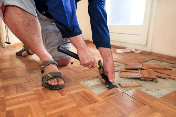 Bouwvakker en constructeur werkt aan renovatie van appartement. Klusjesman is het verwijderen van oude houten parketvloeren met behulp van gele hamer en schrapen gereedschap. Bouwer ontmantelt parketvloer. — Stockfoto