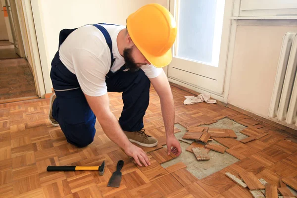 Építőipari munkás és kivitelező dolgozik felújítása lakás. Handyman eltávolítja a régi fa parketta segítségével sárga kalapács és kaparó eszköz. Builder leszerelni parketta padló. — Stock Fotó