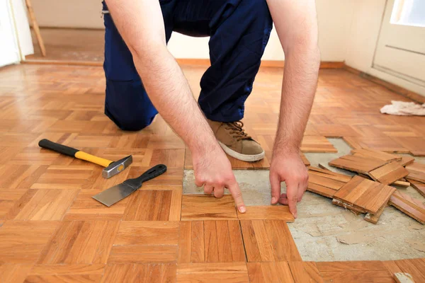 Construction worker and constructor is working on renovation of apartment. Handyman is removing old wooden parquet flooring using yellow hammer and scraping tool. Builder dismantles parquet floor. Royalty Free Stock Images