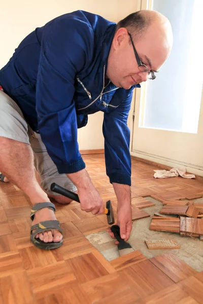 Bouwvakker en constructeur werkt aan renovatie van appartement. Klusjesman is het verwijderen van oude houten parketvloeren met behulp van gele hamer en schrapen gereedschap. Bouwer ontmantelt parketvloer. — Stockfoto