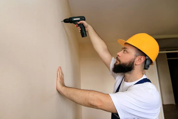 Construction worker and handyman works on renovation of apartment. Builder using electric screwdriver and screwing screw out of wall on construction site. Home renovation concept. Construction tool.