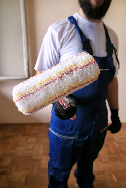 Bouwvakker op bouwplaats met beschermend uniform, bouwgereedschap, uitrusting en instrument. Hij in blauwe overall en houdt verf roller borstel voor muur schilderen in de hand. — Stockfoto