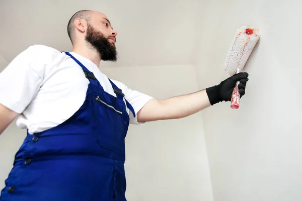 Construction worker doing finish renovation at apartment. Professional painter contractor using white paint roller brush painting of concrete wall room on construction site. Home renovation concept.