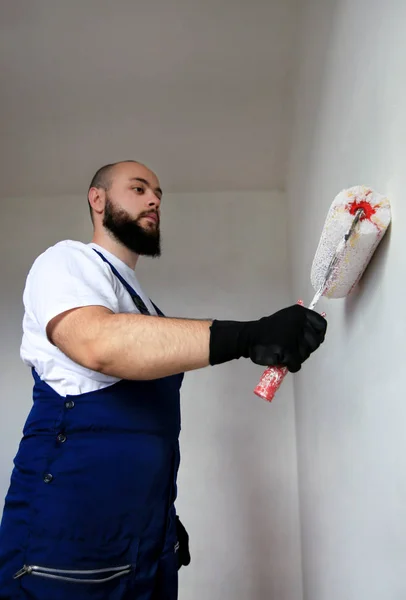 Bouwvakker doet afwerking renovatie in het appartement. Professionele schilder aannemer met behulp van witte verf roller borstel schilderen van beton muur kamer op de bouwplaats. Huisrenovatieconcept. — Stockfoto