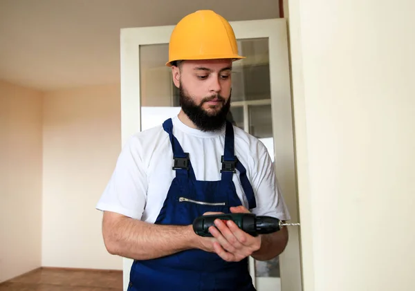 Construction worker and handyman works on renovation of apartment. Builder with electric drill drills nail hole into wooden wall door at construction site. Home renovation concept. Construction tool. Royalty Free Stock Images