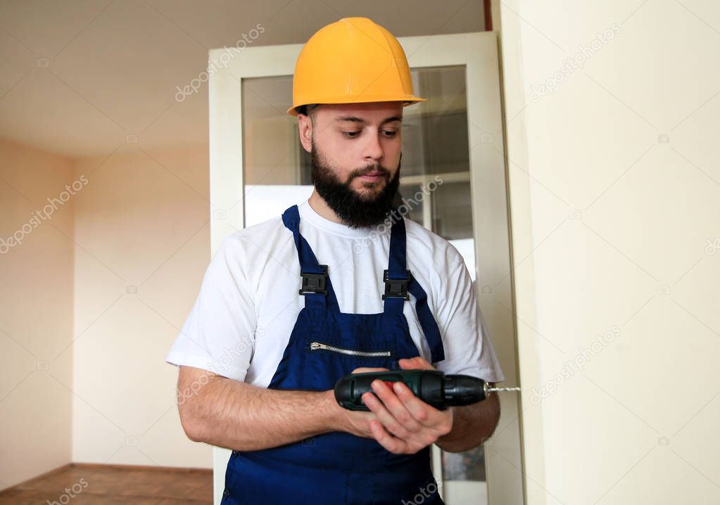 Construction worker and handyman works on renovation of apartment. Builder with electric drill drills nail hole into wooden wall door at construction site. Home renovation concept. Construction tool.