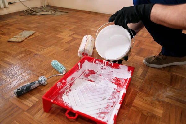 Bouwvakker en klusjesman doen renovatie in appartement. Professionele schilder met zwarte handschoenen giet witte kleur van emmer in rode kom voor het mengen van verf. Huisrenovatieconcept. — Stockfoto