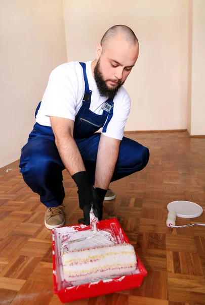 Bouwvakker en klusjesman doen renovatie in appartement. Professionele schilder met handschoenen houdt witte roller borstel in zijn hand en mengt verf in container en rode kom voor kleurmenging. — Stockfoto