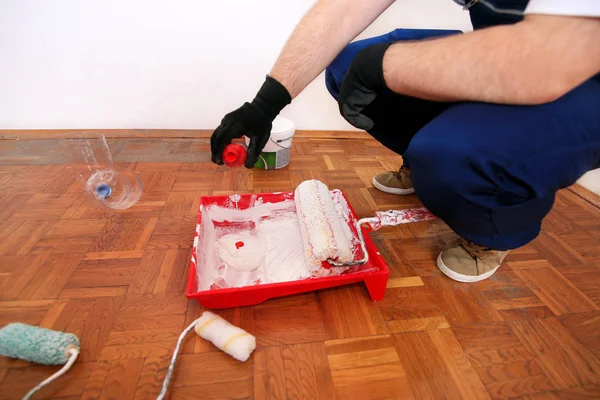 Construction worker doing finish renovation at apartment. Painter with black gloves pours red color pigment for painting wall of room from bottle into bowl for paint mixing. Home renovation concept.