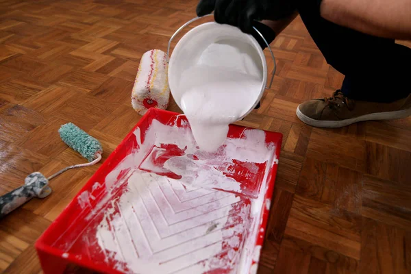 Construction worker and handyman doing finish renovation at apartment. Professional painter with black gloves pours white color from bucket into red bowl for paint mixing. Home renovation concept. Royalty Free Stock Photos