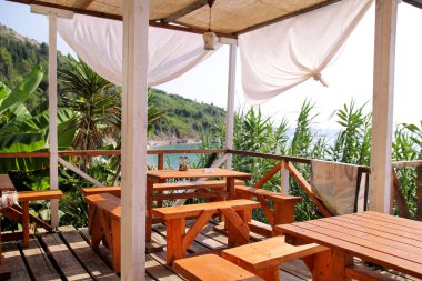 Açık deniz manzaralı deniz kıyısındaki tropik plaj kafe barı. Teras balkonu, deniz kenarındaki ahşap çitli, boş masalı, sandalyeli ve deniz manzaralı tatil beldesi yemek ve içmek için rahat bir yer..