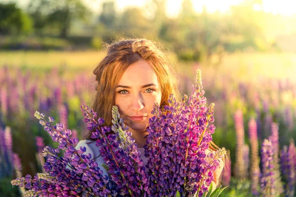 beautiful sexy woman in white sundress with a bouquet in the hands of lupine in field at sunset