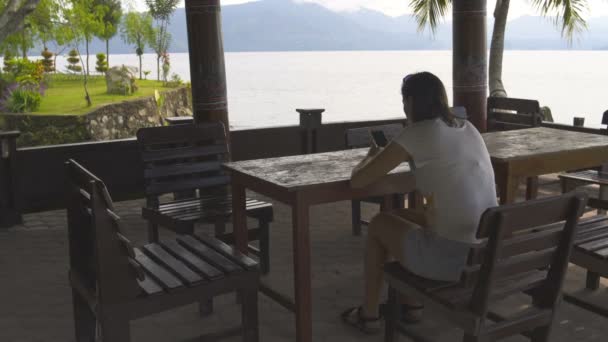 A woman with a smartphone sits at a table overlooking the lake and mountains — Stock Video