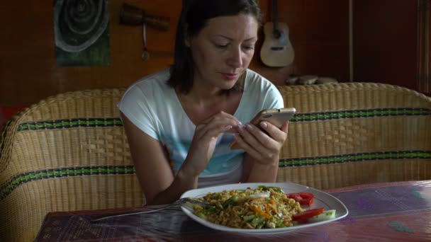 Mujer en un café come Mia Goreng y utiliza el teléfono inteligente — Vídeo de stock