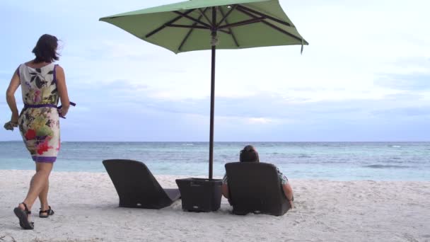 Uomo sdraiato su una poltrona sulla spiaggia. Una donna viene a sdraiarsi su un lettino — Video Stock
