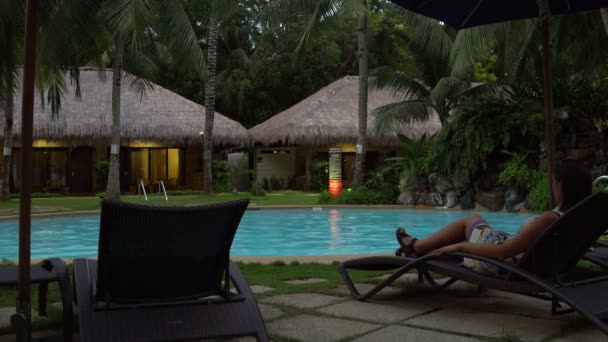 La mujer está acostada en una tumbona junto a la piscina al aire libre por la noche — Vídeo de stock