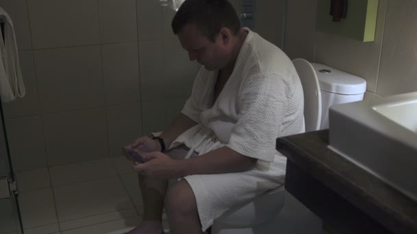 Ein Mann im Kleid sitzt auf der Toilette und benutzt ein Smartphone — Stockvideo