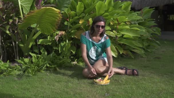Flicka sitter på gräset och äta en mango. I närheten står en skål med frukt — Stockvideo