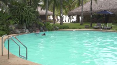 Yapay bir şelale altında bir havuzda bir adam duruyor. Kadın yüzer sırtüstü