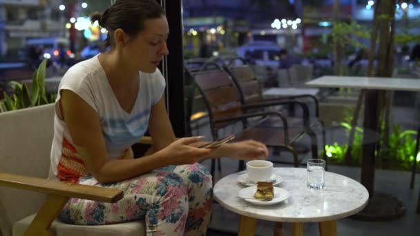 A woman in a cafe with a smartphone drinks coffee and eats a tiramisu cake — Stock Video